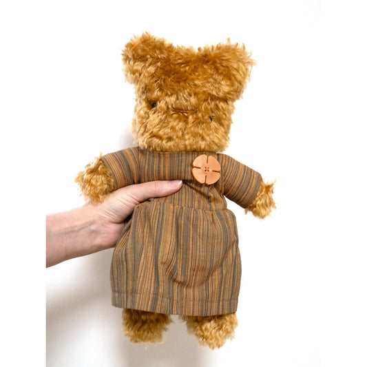 This is Autumn by Bear Bears. Medium-sized teddy named Autumn with curly, medium-long mohair fur that has a soft sheen; the fur is mainly brown with lighter beige highlights. The muzzle is brown and blends with the surrounding fur. Small dark black eyes and a narrow stitched brown nose are visible. The paws and visible paw ends are the same brown fur. The teddy wears a removable striped dress in brown and blue tones with a large round button sewn to the front; the dress fabric shows fine horizontal str...