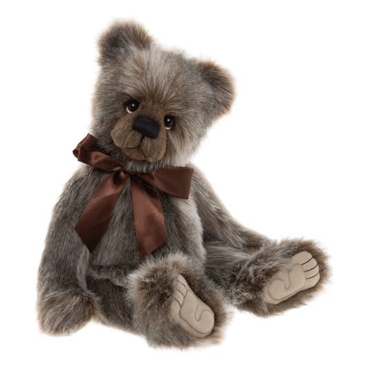 Das ist Ollie, ein Teddy von Charlie Bears. Er sitzt und hat langes, glattes Fell in Grau mit braunen Schattierungen und kleinen weißen Stellen. Die Schnauze ist braun, die Ohreninnenseiten sind grau mit braunen Rändern, die Pfotenballen beige. Die Augen sind dunkelbraun und glänzend, darunter kleine weiße Halbmonde, die Nase ist schwarz und länglich-oval, der Mund aufgestickt. Am Hals sitzt eine große rostbraune Satinschleife, der Ausdruck wirkt sanft und verträumt.