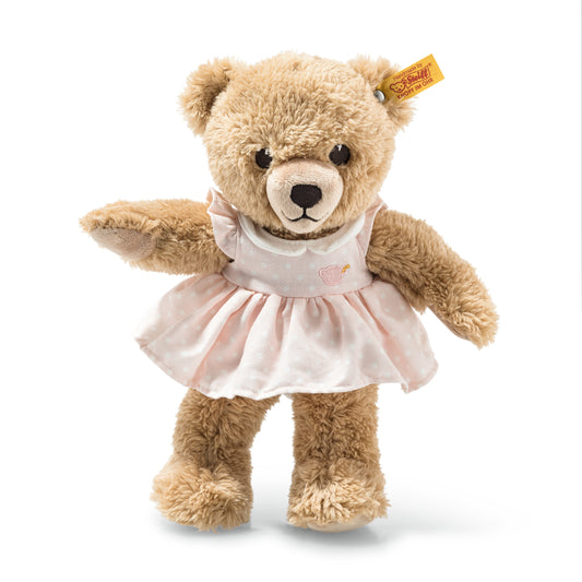 Das ist Sleep Well Bear, ein Teddy von Steiff. Ein mittelgroßer Bär mit langem, leicht zotteligem beigem Fell in weichen Locken und mattem Glanz. Schnauze und Pfotenballen sind heller weiß, die Ohreninnenseiten beige. Die Augen sind schwarz und rund, die Nase schwarz und rundlich, darunter ein gestickter schwarzer Mund. Sie trägt ein abnehmbares rosa Kleid mit weißen Punkten, Stickmotiv am Oberteil, gekraustem Rock und Kragen. Sie sitzt und wirkt ruhig und schläfrig.