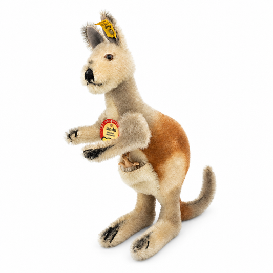 Das ist Linda, ein Känguru von Steiff. Handgefertigt aus Mohair, fest in der Haptik, steht sie aufrecht auf den Hinterbeinen. Kopf, Brust, Beine, Schnauze und Beutel sind beige, Rücken und Hinterteil warm braun, der Schwanz leicht grau. Das Fell ist kurz, fein, etwas zottelig und leicht glänzend. Ohreninnenseiten dunkelgrau, Pfotenspitzen und Krallen schwarz, Augen braun, Nase schwarz und rund. Im Beutel sitzt ein kleines Joey aus Plastik, der Ausdruck wirkt aufmerksam.